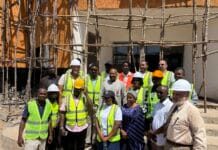 Vice President Intensifies Oversight of BADEA-Funded Projects at Prince of Wales & Milton Margai, Pushes for Timely Completion Group of construction workers in high-visibility vests and hard hats posing in front of a building under wooden scaffolding at a construction site.