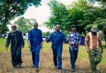 Agriculture Minister Inspects Magburaka Ahead of World Food Day, Feed Salone Anniversary Five men walk together through a dry grassy field with trees in the background, wearing blue and patterned shirts.