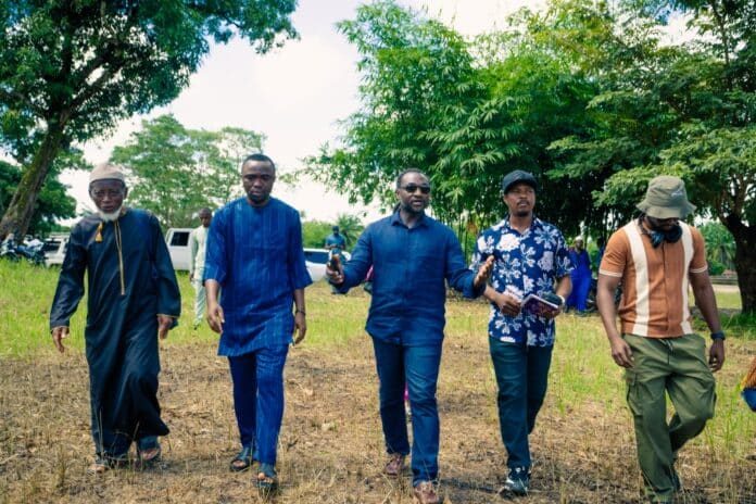 Five men walk together through a dry grassy field with trees in the background, wearing blue and patterned shirts.