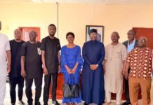 Employers Federation Pledges Cooperation with Government on Labour Reforms Group of eleven adults posing in a line in a brightly lit room, wearing traditional African attire; a framed photo hangs on the wall behind them.