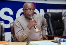 Government Gazettes New Districts, City Councils, Rural Councils, Chiefdoms and Villages Man in glasses and a beige traditional shirt speaks into a handheld microphone at a conference table, with papers and a water bottle in view.