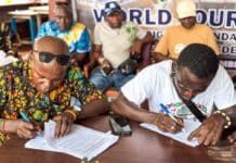 Eight Peninsula Communities Sign Declaration to Protect Beaches, Boost Sustainable Tourism Two men sign documents at a wooden table during a meeting; one in a colorful patterned shirt with sunglasses, the other in a white T‑shirt; others sit in the background.