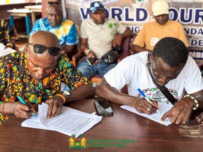 Two men sign documents at a wooden table during a meeting; one in a colorful patterned shirt with sunglasses, the other in a white T‑shirt; others sit in the background.