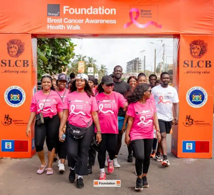 Group of people wearing pink Breast Cancer Awareness shirts walking under an orange Foundation arch at a charity health walk.