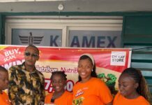 Alice George Bags NLe17,500 as First Grand Prize Winner in Nutribom “Buy, Scan and Win” Promotion Five people stand outside EM Kay Store, four women in orange event shirts and a man in a patterned shirt, handing over a stack of cash alongside a large promotional check to celebrate a prize.