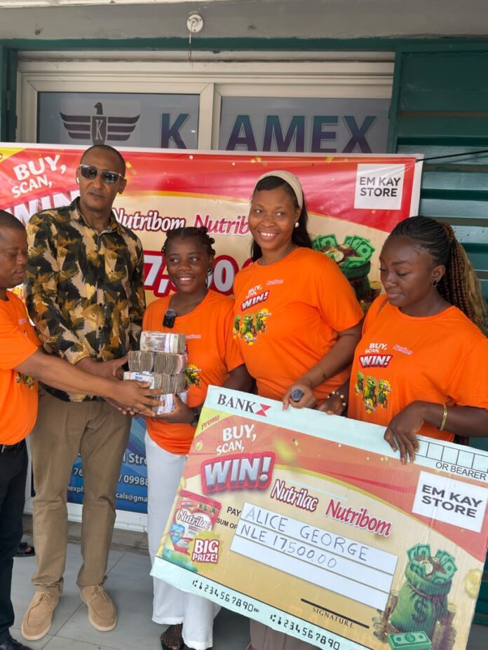 Five people stand outside EM Kay Store, four women in orange event shirts and a man in a patterned shirt, handing over a stack of cash alongside a large promotional check to celebrate a prize.