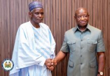 President Bio Receives Alhaji Amadu Juldeh Sowe as New Tabital Pulaaku International President Two men shake hands in front of a wood-paneled wall; one wears traditional white agbada, the other a green official uniform with a small flag pin, a presidential seal visible in the corner.