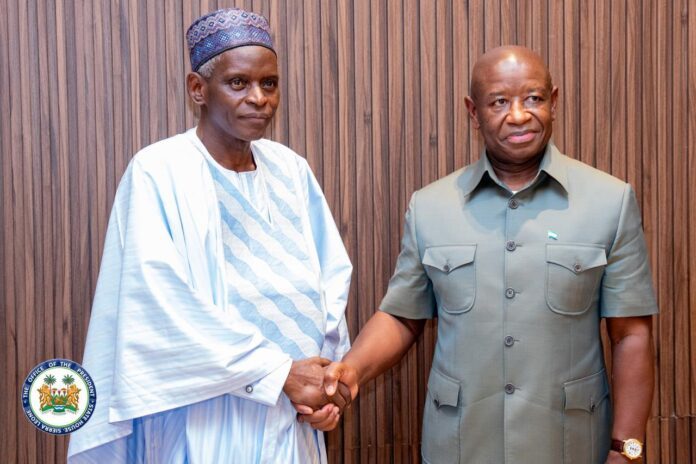 Two men shake hands in front of a wood-paneled wall; one wears traditional white agbada, the other a green official uniform with a small flag pin, a presidential seal visible in the corner.