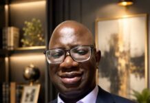 Ahead of 2028 Elections, Sorie Fofana Warns SLPP Against Internal Division Man in a navy suit and light purple shirt wearing glasses, standing in a modern office setting with shelves and framed art behind him.