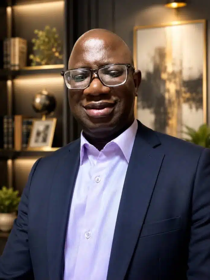 Man in a navy suit and light purple shirt wearing glasses, standing in a modern office setting with shelves and framed art behind him.