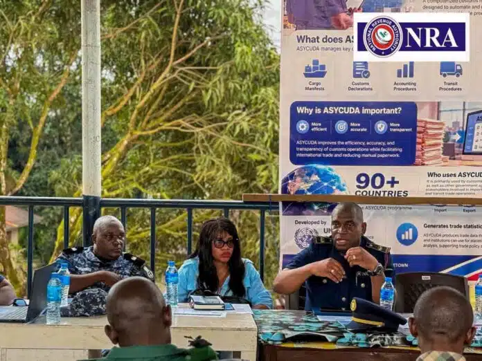 Panel of officials seated at a long table outdoors, with water bottles and papers, in front of a large poster about ASYCUDA.