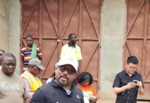 SLRA Advances 97km Magburaka-Bumbuna-Sambaia Bendugu Road Project with Pre-Bid Site Visit Speaker wearing a white cap, navy polo, and cargo pants addresses a small crowd outdoors near a rusty gate area, holding a rolled paper or document.