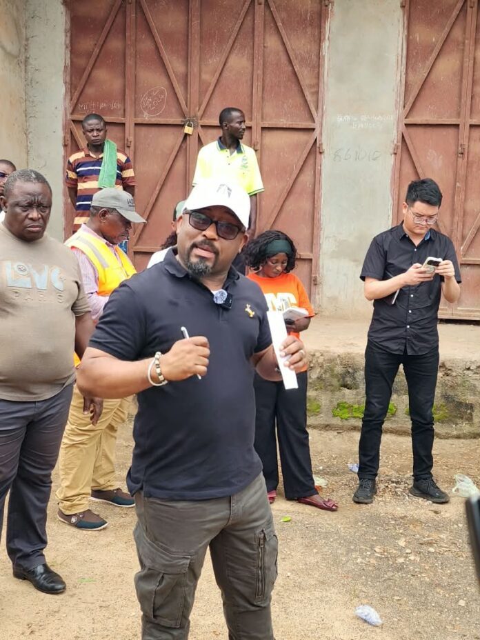 Speaker wearing a white cap, navy polo, and cargo pants addresses a small crowd outdoors near a rusty gate area, holding a rolled paper or document.