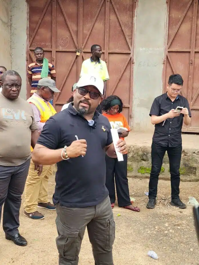 Speaker wearing a white cap, navy polo, and cargo pants addresses a small crowd outdoors near a rusty gate area, holding a rolled paper or document.