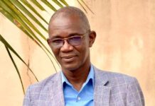 MoCTI Vindicated as Procurement Review Panel Dismisses TMM’s Complaint Over Digital Transformation Project Portrait of a man in a blue button-down shirt and gray plaid blazer, standing outdoors with a palm plant nearby.