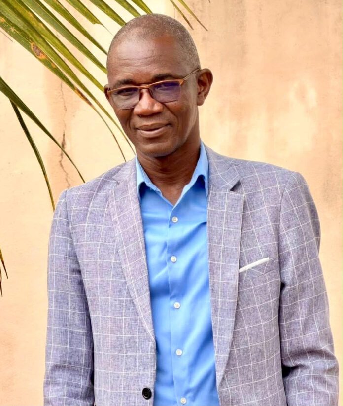Portrait of a man in a blue button-down shirt and gray plaid blazer, standing outdoors with a palm plant nearby.