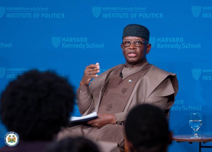 African man in traditional brown attire, speaking with a microphone against a blue Harvard Kennedy School Institute of Politics backdrop, audience in foreground