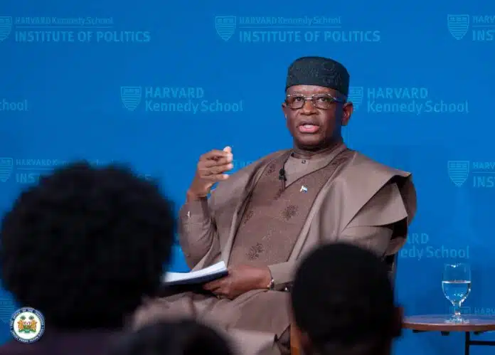 African man in traditional brown attire, speaking with a microphone against a blue Harvard Kennedy School Institute of Politics backdrop, audience in foreground