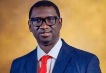 Dr. Ibrahim Bangura Calls for Fair Supreme Court Hearing on Constitution Amendment Bill Professional portrait of a young man wearing a dark blue suit, white shirt, and red tie, with black glasses, set against a warm gold background.