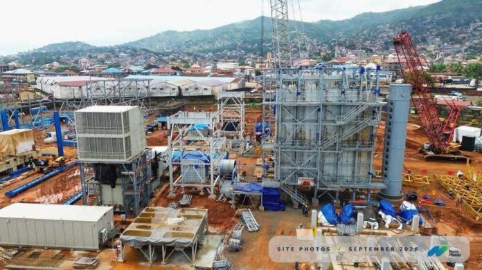 Construction site with tall steel scaffolding, machinery, and a red crane, set against a rural hillside backdrop.