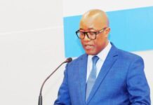 Foreign Minister Unveils Sierra Leone’s Diplomatic Priorities at Maiden Briefing Man in a blue suit and glasses speaks into a microphone at a podium with a blue backdrop.