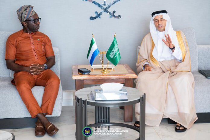 Two men sit on opposite couches in a formal meeting, with Sierra Leone and Saudi flags on a table between them.