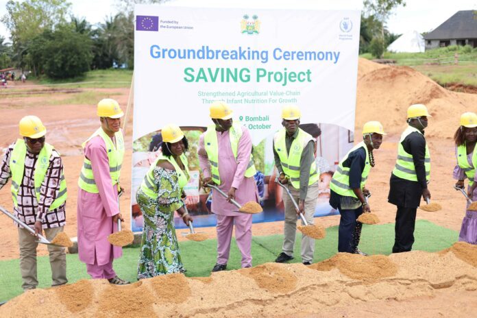 Group of people in yellow hard hats and safety vests shoveling sand at a groundbreaking ceremony in front of a banner reading 'Groundbreaking Ceremony SAVING Project'