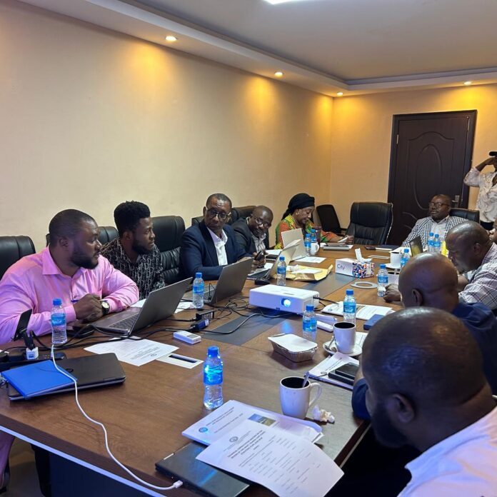 A group of professionals sit around a conference table with laptops, papers, and water bottles during a meeting.