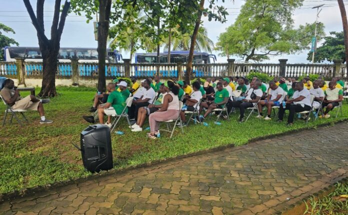 Outdoor community meeting: a speaker sits left addressing a seated audience under trees, many wearing green shirts; buses parked in the background.