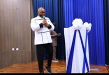 NatCA Champions Digital Safety as Government Intensifies Crackdown on Mobile Money Fraud and Online Scams Man in a white jacket speaking into a microphone on a stage with blue curtains and white/blue draped decorations.
