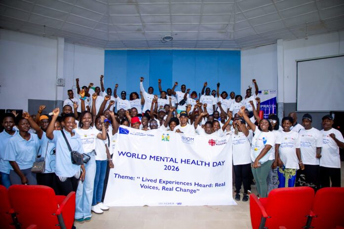 Large group of diverse people in a hall, posing with a banner that reads World Mental Health Day 2026 and the theme Lived Experiences Heard: Real Voices, Real Change, raising their fists in celebration.
