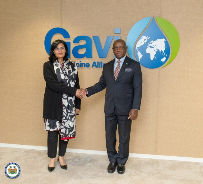 Two formally dressed individuals shake hands in front of the Gavi Vaccine Alliance logo backdrop.