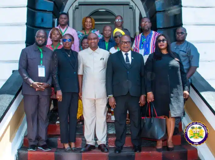 Group of professionals in business attire posing on a staircase outside a building, with the Judiciary of Sierra Leone emblem visible in the lower right corner.