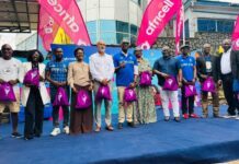 Africell Unveils 2026 Freetown Marathon with Historic 42.2KM Race and Increased Cash Prizes Diverse group of about 12 people standing in a line on a blue stage outdoors, each holding a pink drawstring bag, with bright pink banners in the background, posing for a promotional event photo.