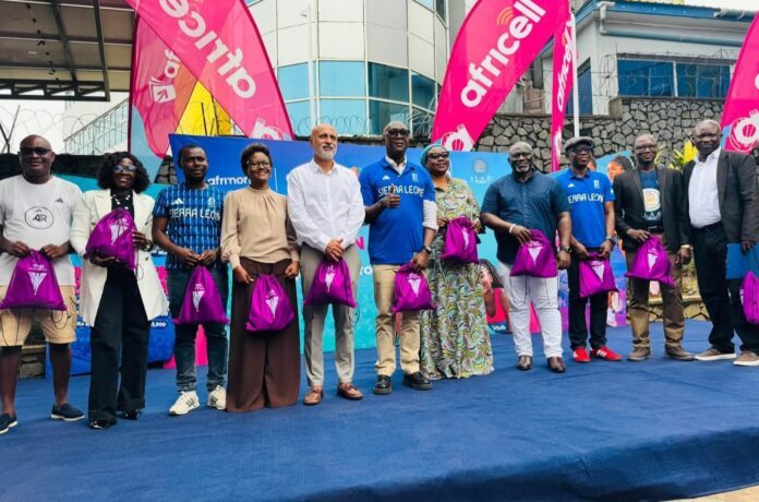 Diverse group of about 12 people standing in a line on a blue stage outdoors, each holding a pink drawstring bag, with bright pink banners in the background, posing for a promotional event photo.