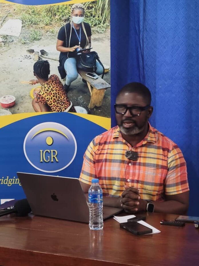 Man in a plaid orange shirt sits at a table with a laptop, microphone, phone, and water bottle during a presentation; banner with IGR logo behind him.