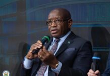 President Bio Calls for Early Intervention and Stronger Institutions to Prevent Conflict Bald man in a dark suit and patterned tie speaks into a handheld microphone on a panel, with a blue backdrop behind him and a small circular logo in the corner.