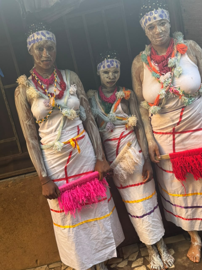 Three people covered in white body paint pose together in traditional white robes decorated with colorful tassels and beads for a cultural festival.