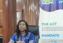 ICPNC Prepares to Host National Unity and Social Cohesion Conference Woman in a blue patterned dress sitting at a desk, speaking, with papers and a water bottle nearby; banner behind reads 'THE ACT' and 'MANDATE'.