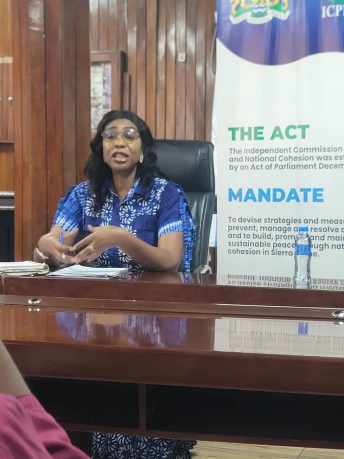 Woman in a blue patterned dress sitting at a desk, speaking, with papers and a water bottle nearby; banner behind reads 'THE ACT' and 'MANDATE'.