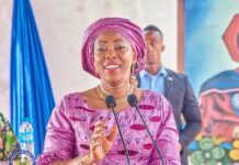 Every Story Is Unique, Every Journey Matters: A Call to Accelerate Action on Women’s Cancers in Africa Woman in purple traditional attire and headwrap speaks at a two-microphone podium, smiling at a formal event with a colorful backdrop behind her.