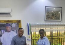 Orange Goes the Extra Mile to Appreciate Customers, Reaffirms Commitment to Service Excellence During Customer Service Week 2026 Two people in a bright office exchange an orange donation box, smiling at the camera with framed portraits and a leather sofa in view.