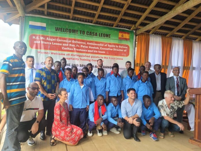 Group of students in blue uniforms and adults posing on a stage under a green banner that reads 'Welcome to Casa Leone' with Sierra Leone and Spain flags nearby, likely a welcome event.
