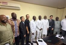 Labour Minister, IOM Strengthen Partnership to Protect Sierra Leonean Migrant Workers A diverse group of men standing in a line in a meeting room, posing for a group photo around a desk.