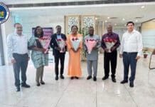 MoH Delegation Visits Allengers Medical Systems in India to Strengthen Hospital Equipment Capacity Seven people stand in a bright lobby, each holding flower bouquets; Ministry of Health logo is visible in the top-left corner.