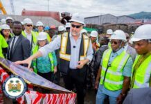 President Bio Inspects US$417 Million Nant Power Project as Sierra Leone Targets Major Electricity Boost in 2027 Senior official in a white hard hat and reflective vest explains a project to a diverse group at an outdoor construction site, with a government seal visible on a barrier nearby.