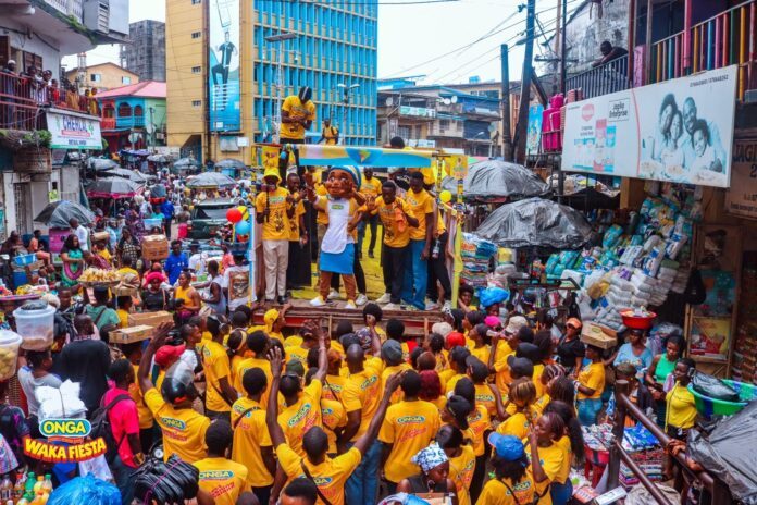 Crowd in a busy market street cheering at a yellow-clad parade with a cartoon mascot on a float.