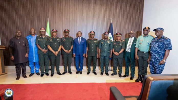 Group of military officers in green uniforms standing with formally dressed civilians in a line, flag backdrop and red carpet.