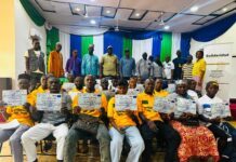 80 Cashew Farmers Trained to Extend Agricultural Knowledge to 300 Others Group of people seated and standing on a stage, proudly holding certificates at a ceremony in a hall with blue, green, and white drapes behind them.