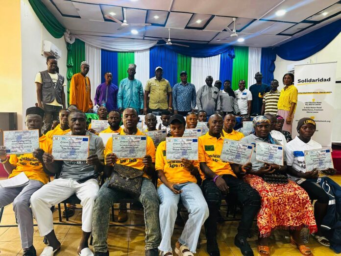 Group of people seated and standing on a stage, proudly holding certificates at a ceremony in a hall with blue, green, and white drapes behind them.
